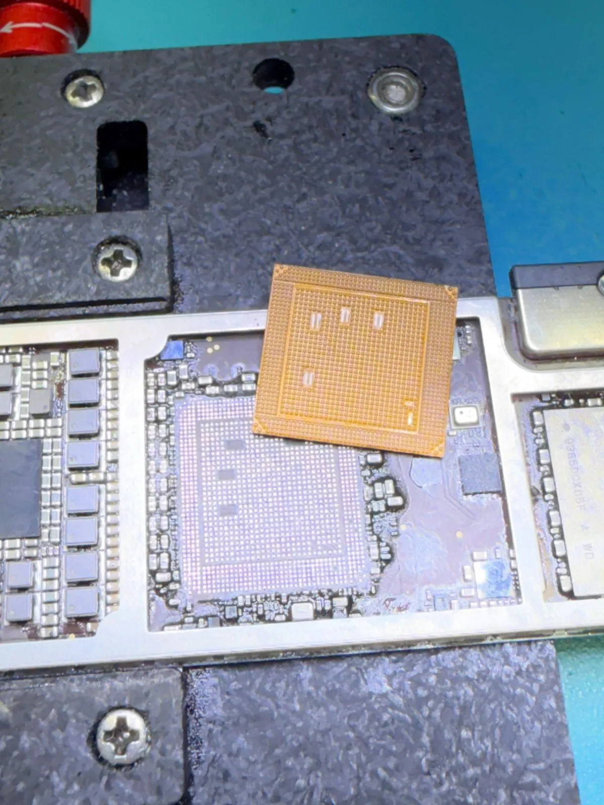 A logic board clamped in a repair fixture with its main chip lifted and resting beside the pads.